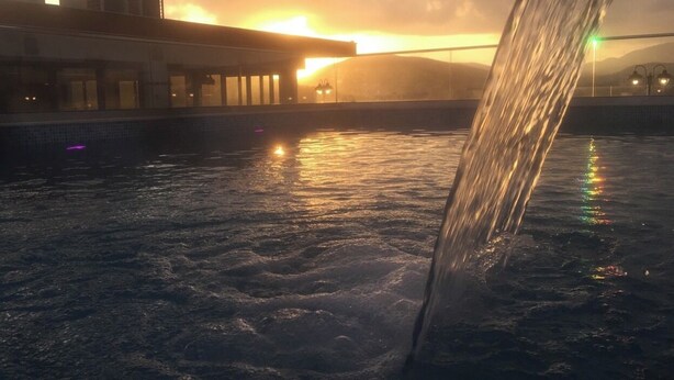 A pool with a waterfall feature, surrounded by a building with large windows and a view of a sunset. - Hotel Evropa (Ulcinj)