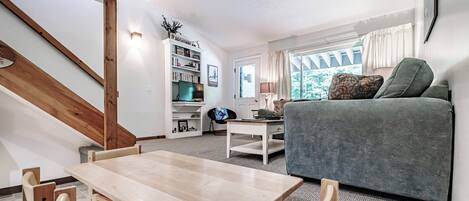 A cozy living room with a wooden dining table, a grey sofa, and a bookshelf.
