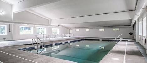 Una piscina interior con agua cristalina, bordes azulejados y suelo de hormigón. Hay sillas y mesas cerca de la piscina, y ventanas que dejan entrar luz natural.
