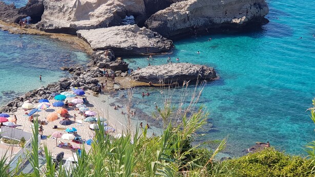 A rocky beach with clear blue water, umbrellas, and people swimming.