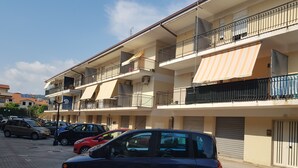 A multi-story residential building with balconies, a parking area, and a clear sky.