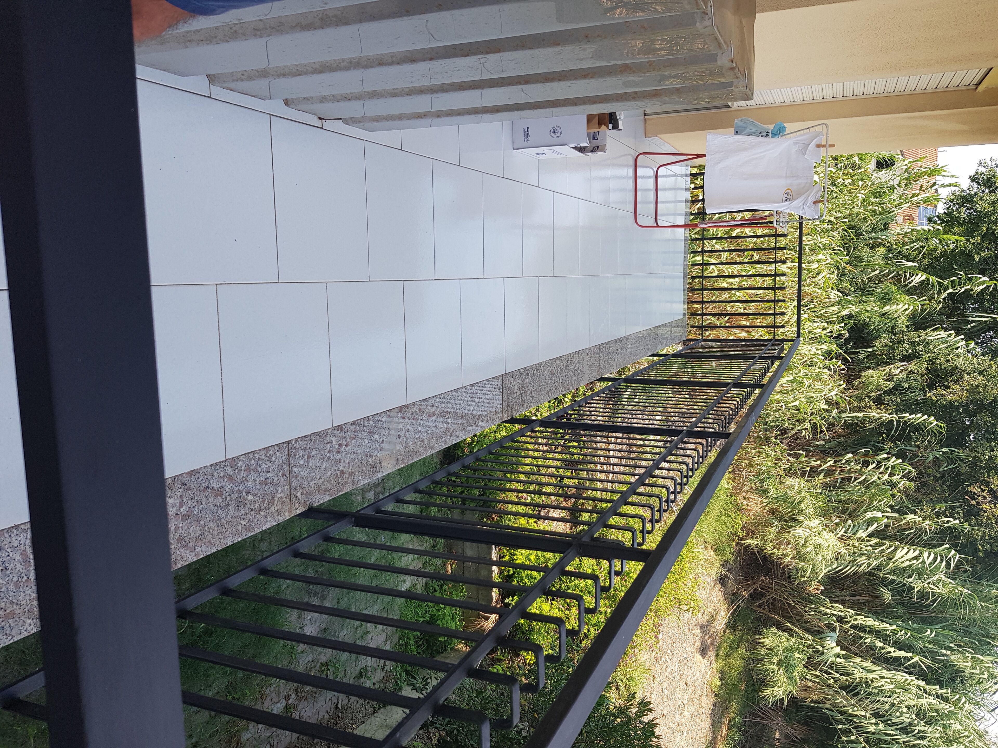 Un balcon doté d’une rambarde en métal, d’un sol carrelé de couleur blanche et offrant une vue sur la verdure et les bâtiments.
