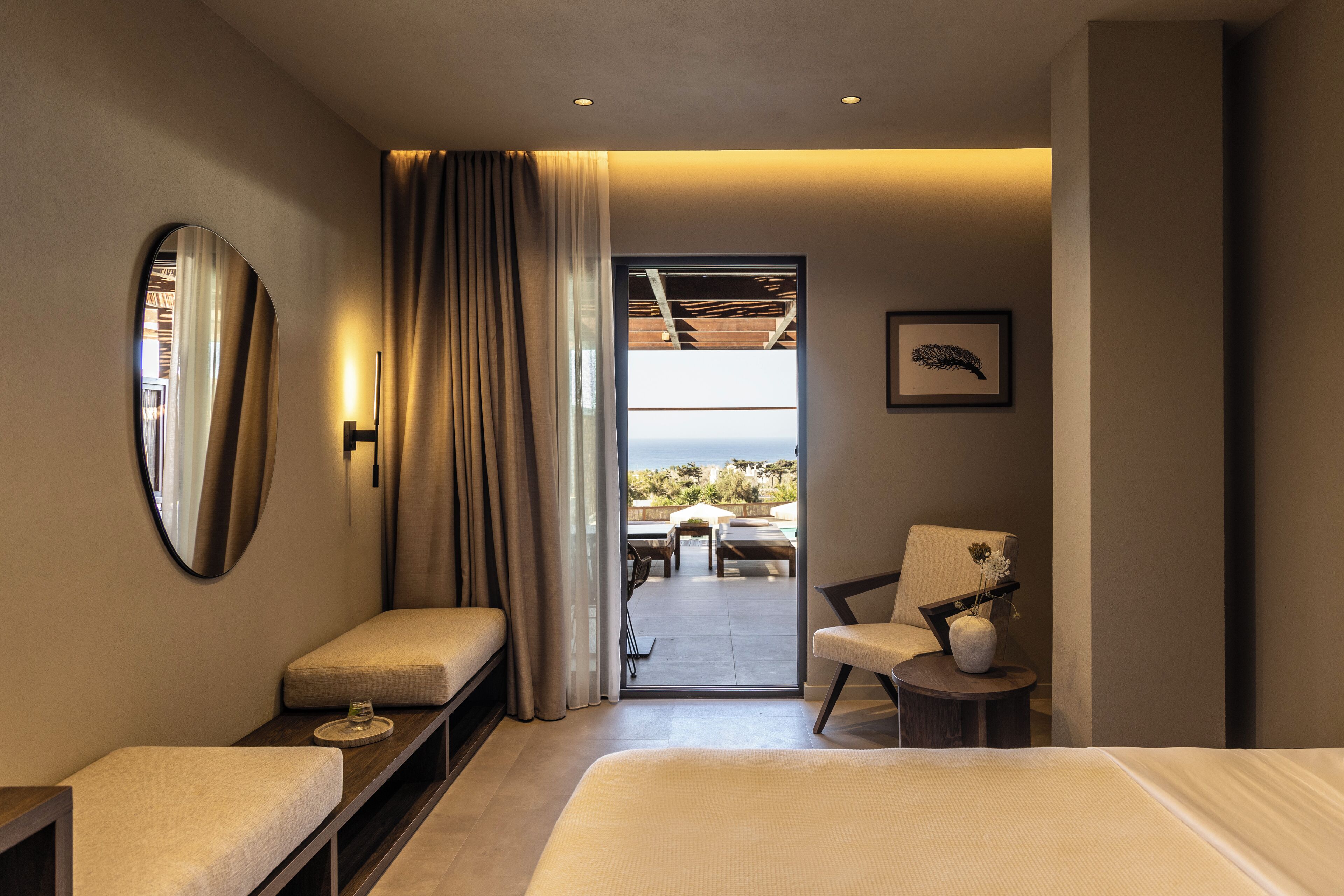 A modern hotel room with a large bed, a seating area with a chair and table, a small side table, a mirror, and a view of the outdoors through a glass door.