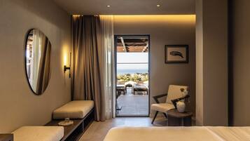 A modern hotel room with a large bed, a seating area with a chair and table, a small side table, a mirror, and a view of the outdoors through a glass door.