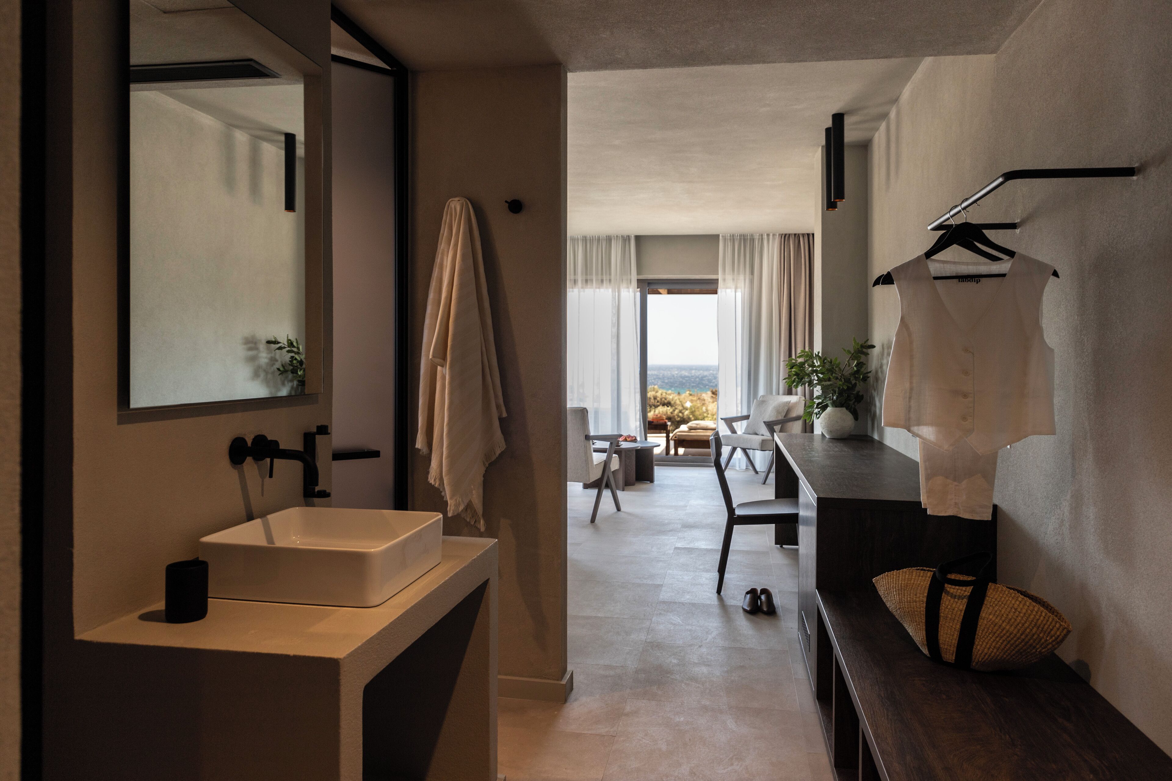 A modern bathroom with a large mirror, a sink, and a towel rack. There is a view of a dining area with a table and chairs in the background.