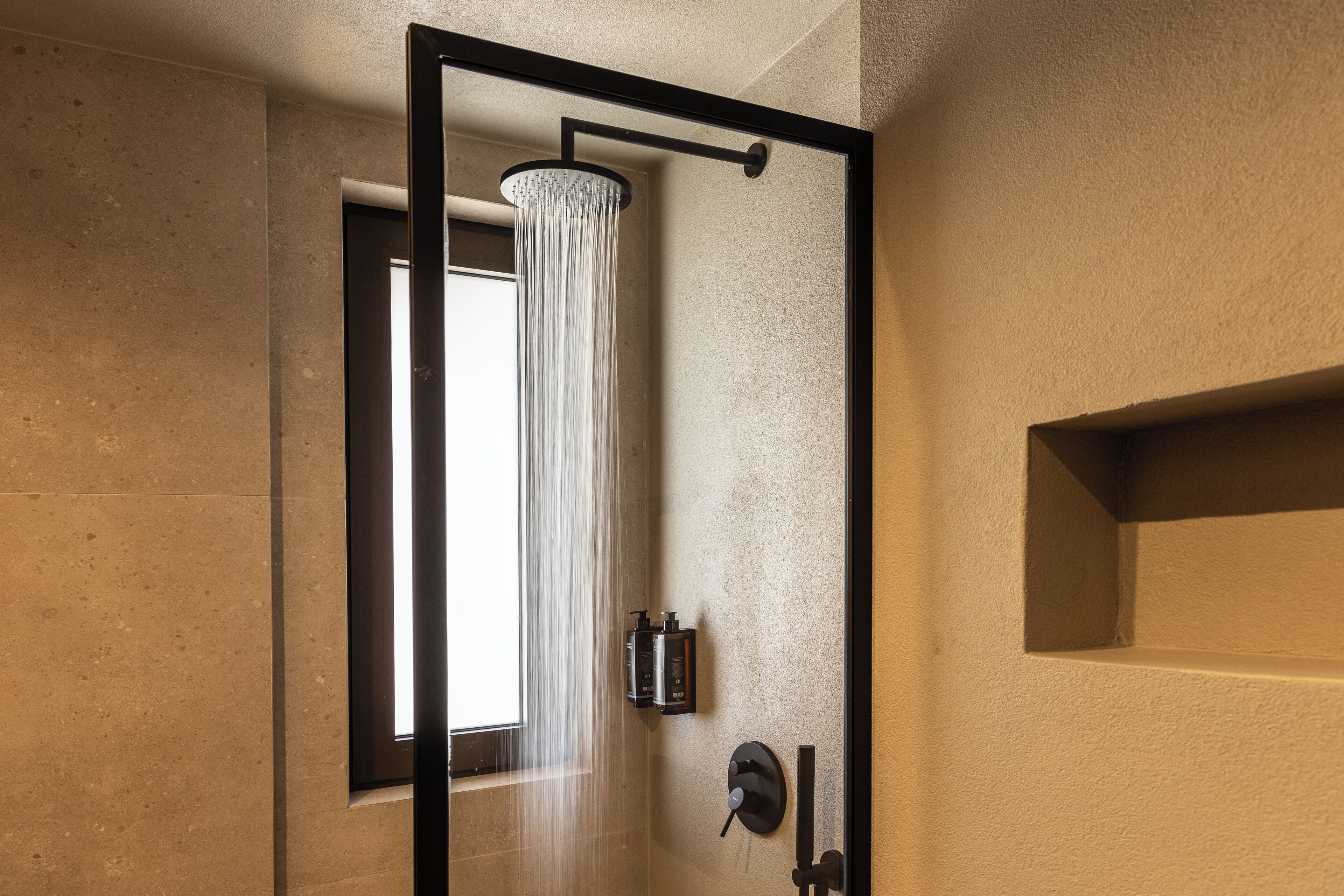 Un baño moderno con ducha de vidrio, una ventana visible y dispensador de jabón en la pared.