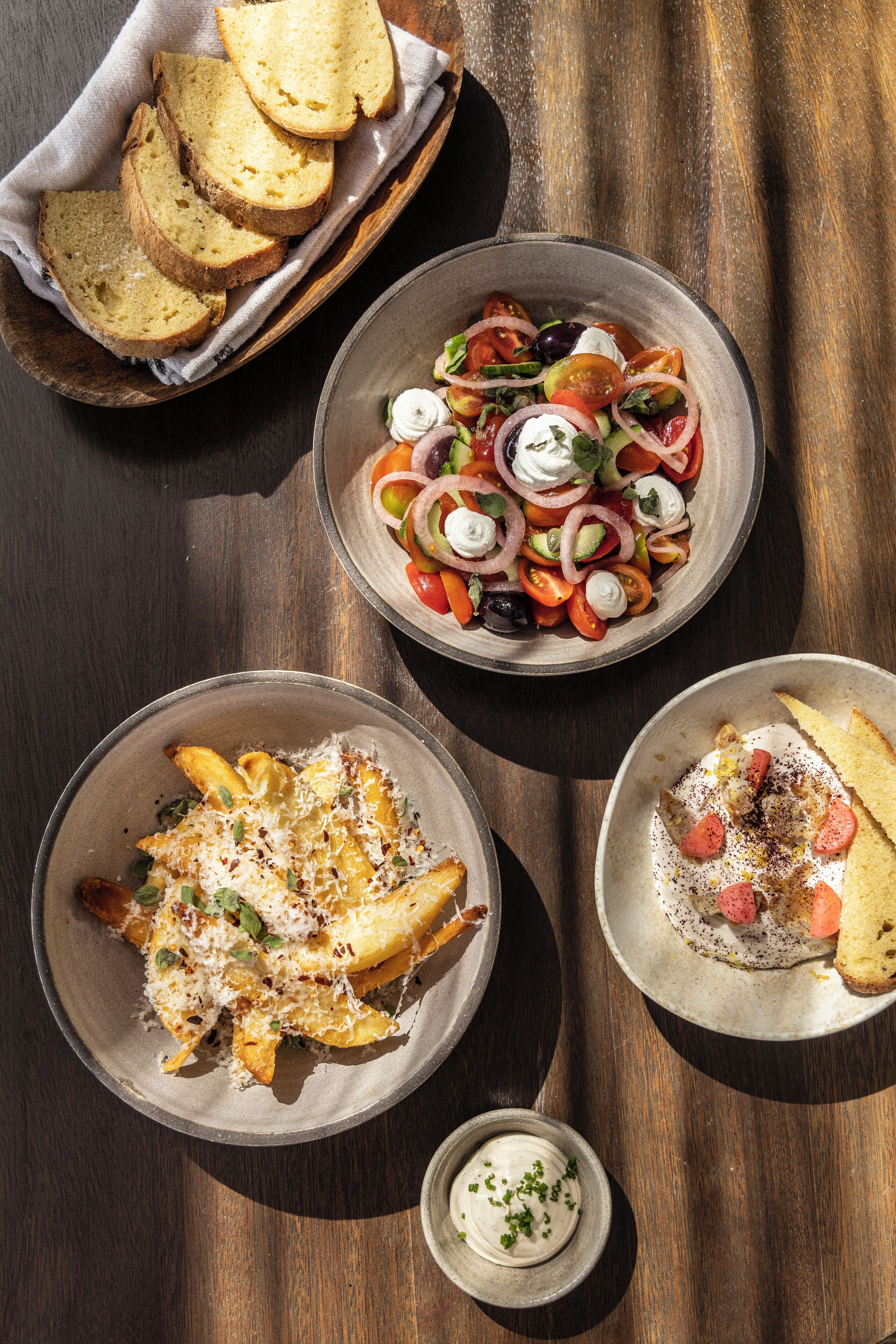 A wooden table with four dishes: a salad, a dish with fries, a bread basket, and a small bowl of sauce.