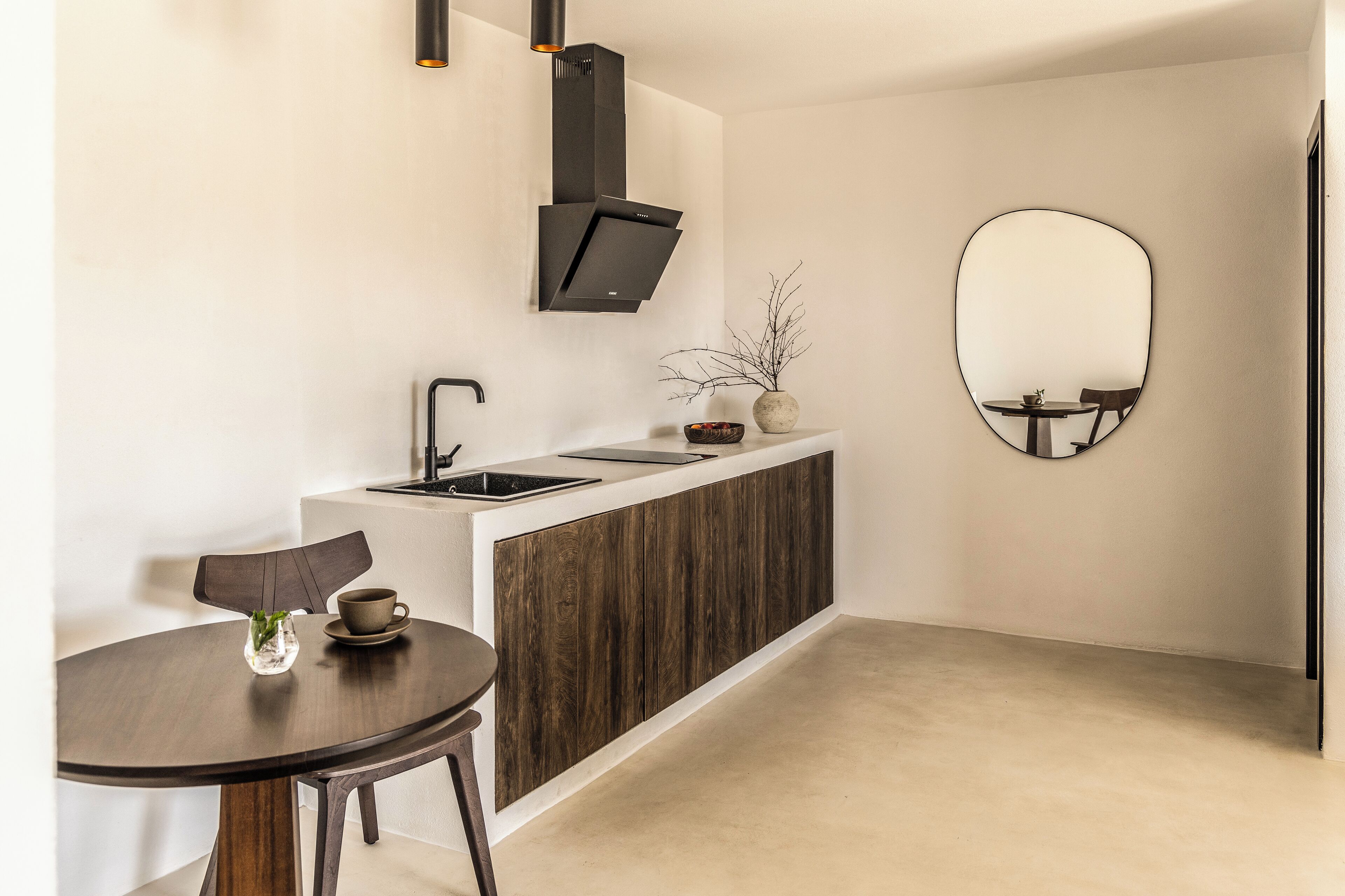 A modern kitchen with a built-in stove, a sink, and a wooden cabinet. There is a round mirror on the wall and a small table with a cup and saucer.