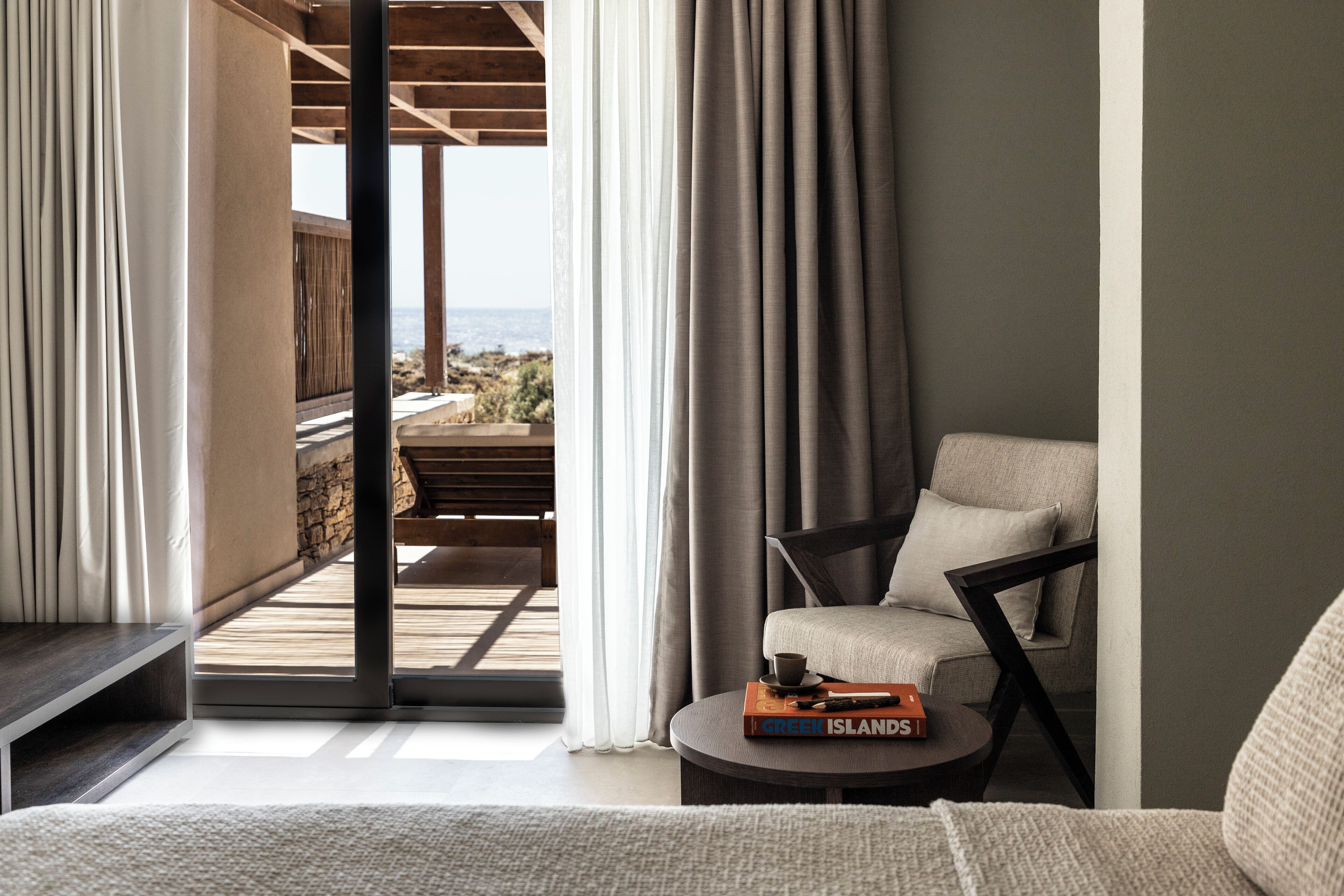 A modern hotel room with a view of the sea, a comfortable sofa, a small table with books, and a balcony with a wooden deck.