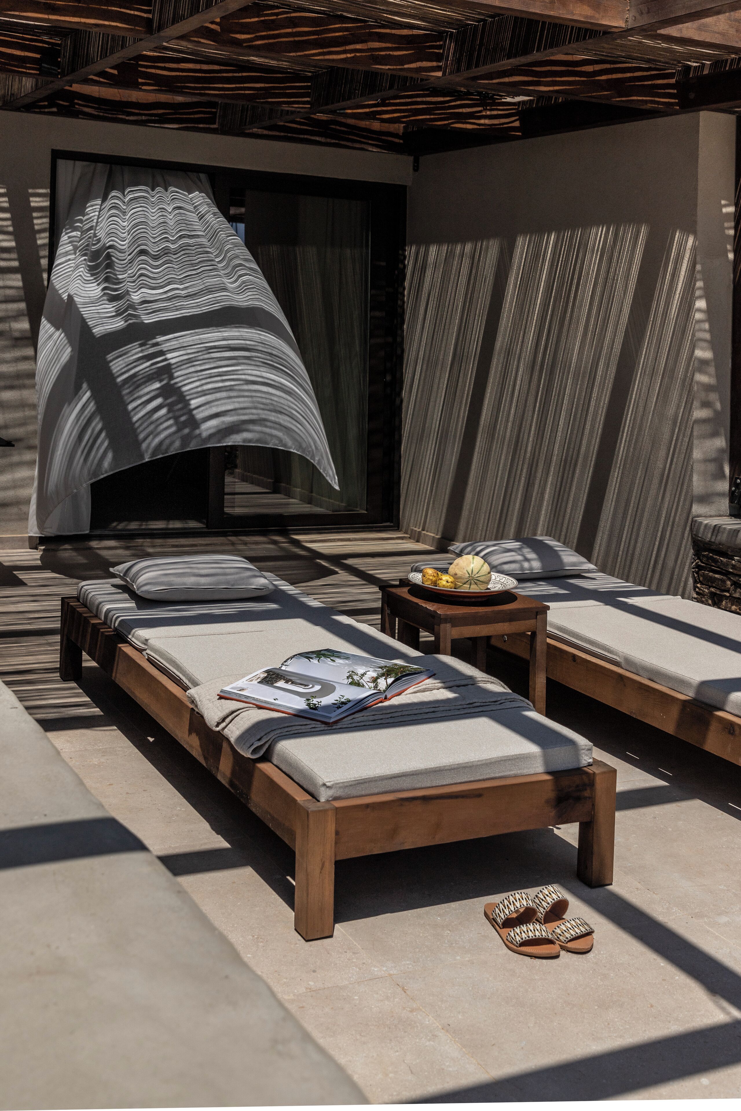 A modern outdoor lounge area with wooden loungers, a small table with a bowl of fruit, and a large abstract artwork on the wall.