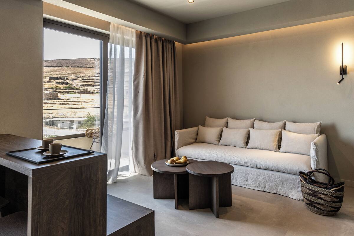 A modern hotel room with a large window, a sofa, a small round table with a tray of fruits, and a dark wooden desk.