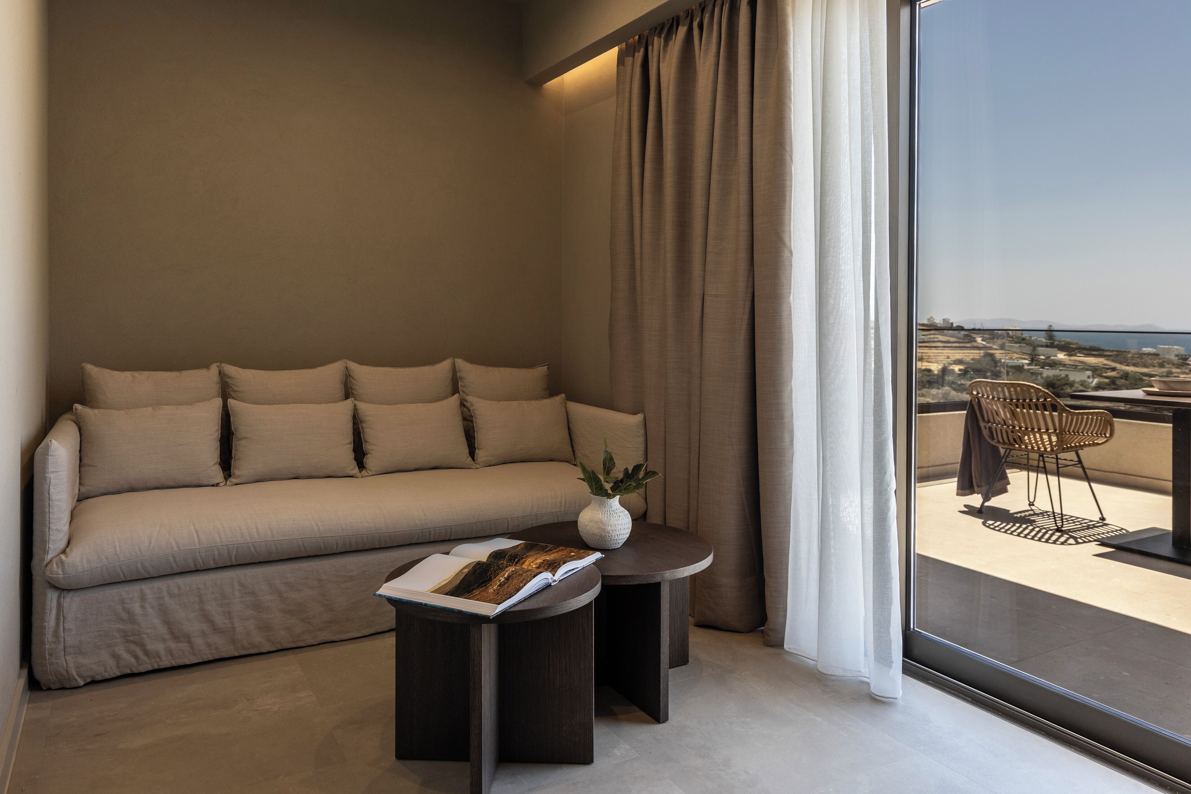 A modern living room with a beige sofa, a small side table with a book, and a view of a balcony with a chair and table.
