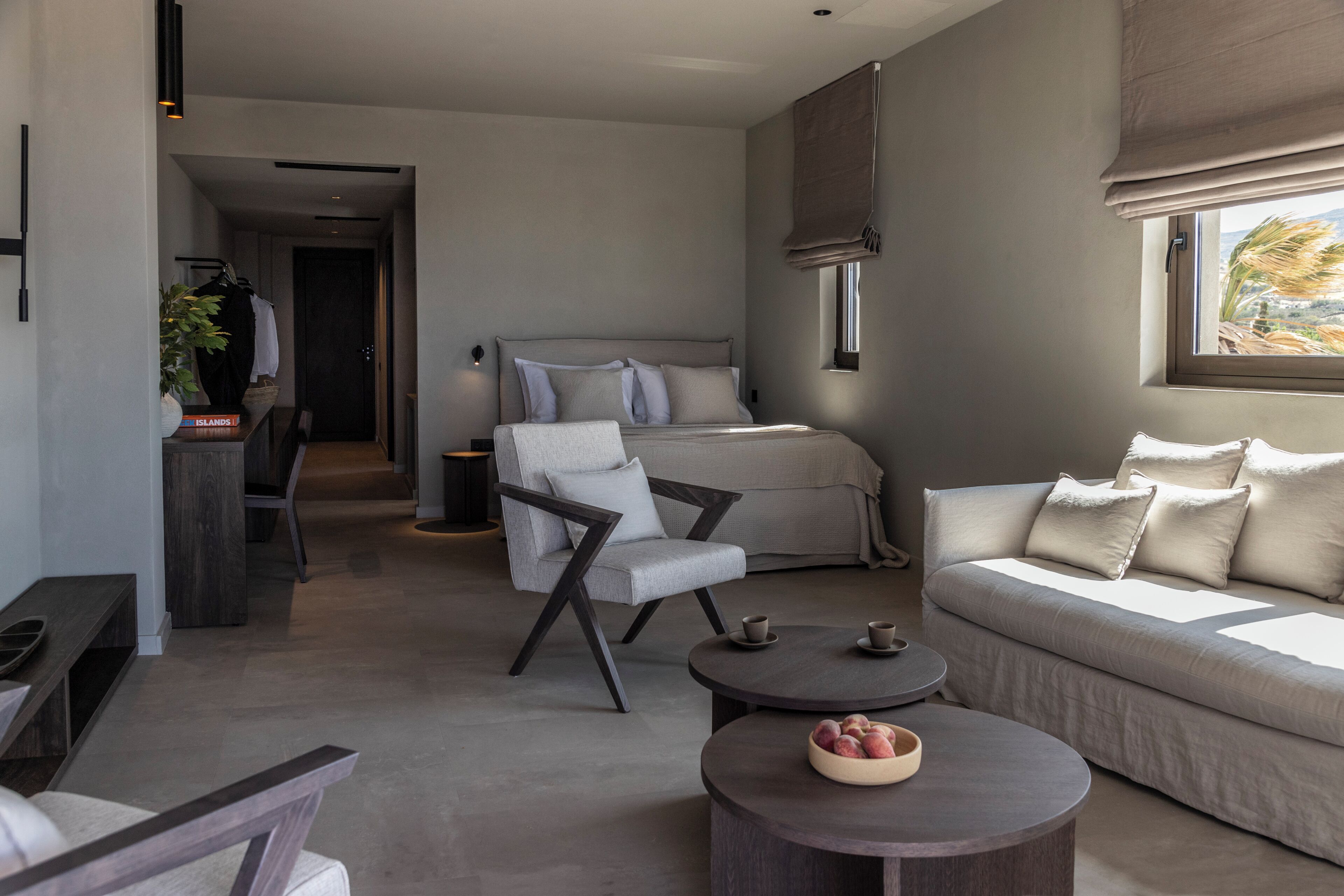 A modern, minimalist living room with a sofa, armchair, and coffee table, a bed with a headboard, and a window with a view.