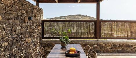 A wooden table with a bowl of fruit and a potted plant, surrounded by wicker chairs, set against a stone wall and wooden ceiling, overlooking a hilly landscape.