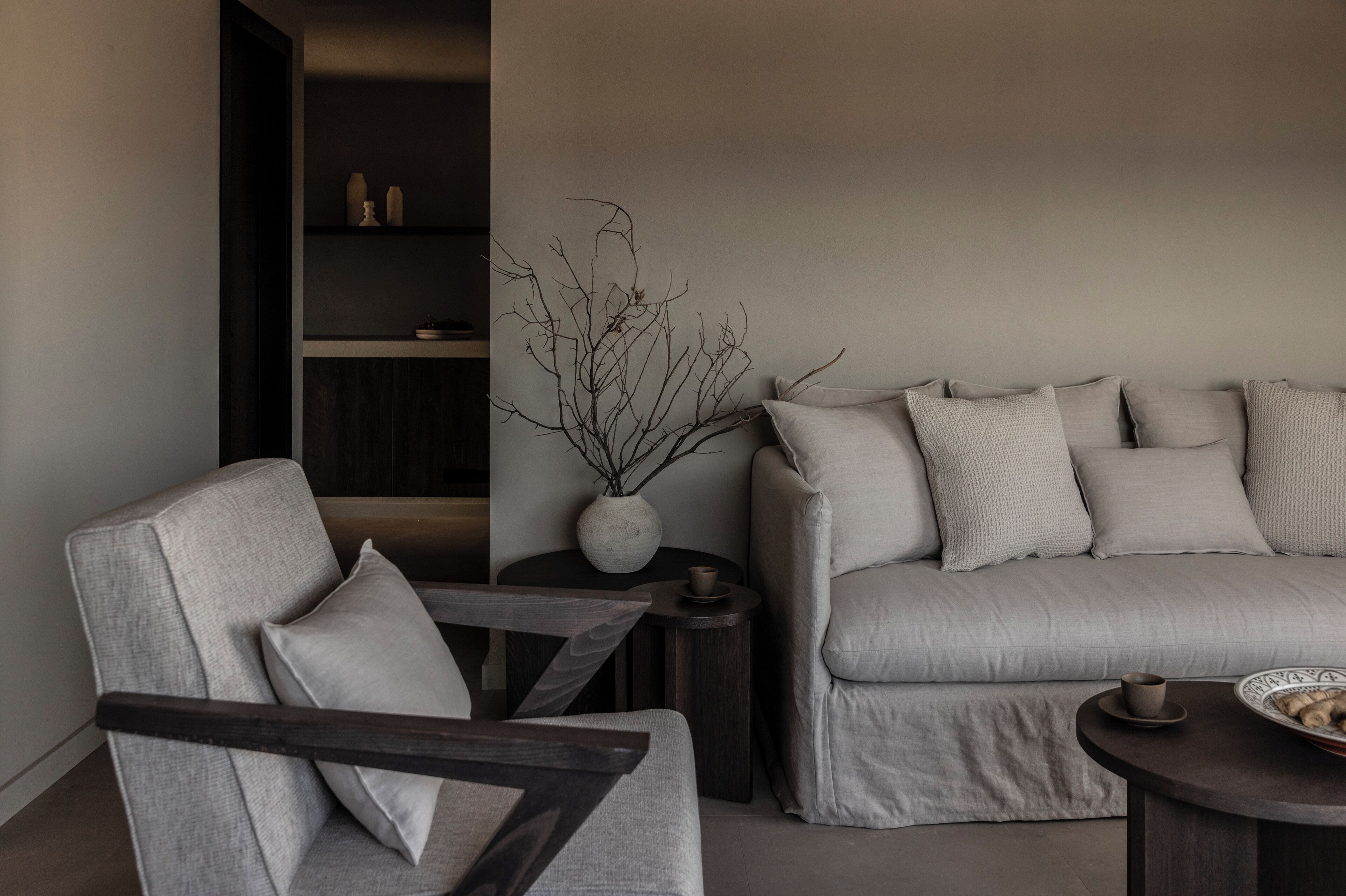 A modern living room with a grey sofa, a matching armchair, a small round table with a cup and saucer, and a vase with branches on a side table.