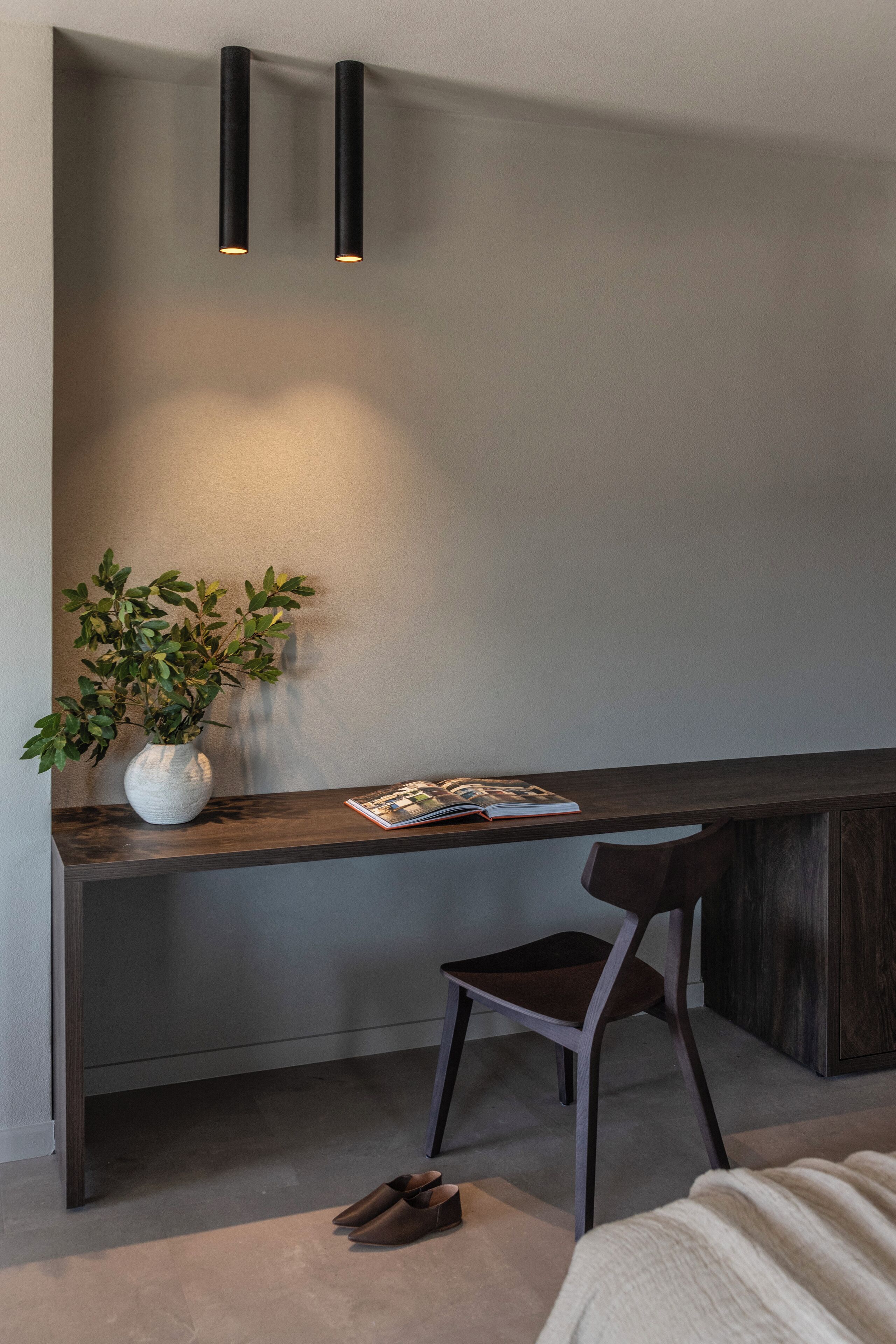 A modern room with a dark wooden desk, a white vase with green plants, a magazine, and a chair.