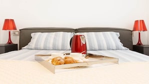 Ein ordentlich bezogenes Bett mit gestreiften Kissen, ein Tablett mit Croissants und einer Kaffeekanne sowie rote Lampen auf beiden Seiten.
