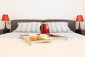 Una cama bien hecha con almohadas a rayas, una bandeja con cruasanes y una cafetera, y lámparas rojas a cada lado.