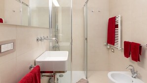 Ein Badezimmer mit einer Glaskabine zur Dusche, einem weißen Waschbecken und einem Handtuchhalter mit roten Handtüchern.