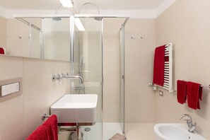 Un baño con ducha de mampara de vidrio, un lavamanos blanco y un toallero con toallas rojas.