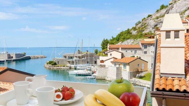 Ein Tablett mit Obst und Tassen auf einem Balkon mit Blick auf eine Marina, in der Boote und Gebäude zu sehen sind.