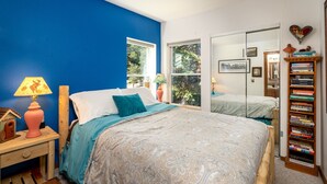 A bedroom with a large bed, a nightstand with a lamp, a bookshelf, and a window with a view of trees.