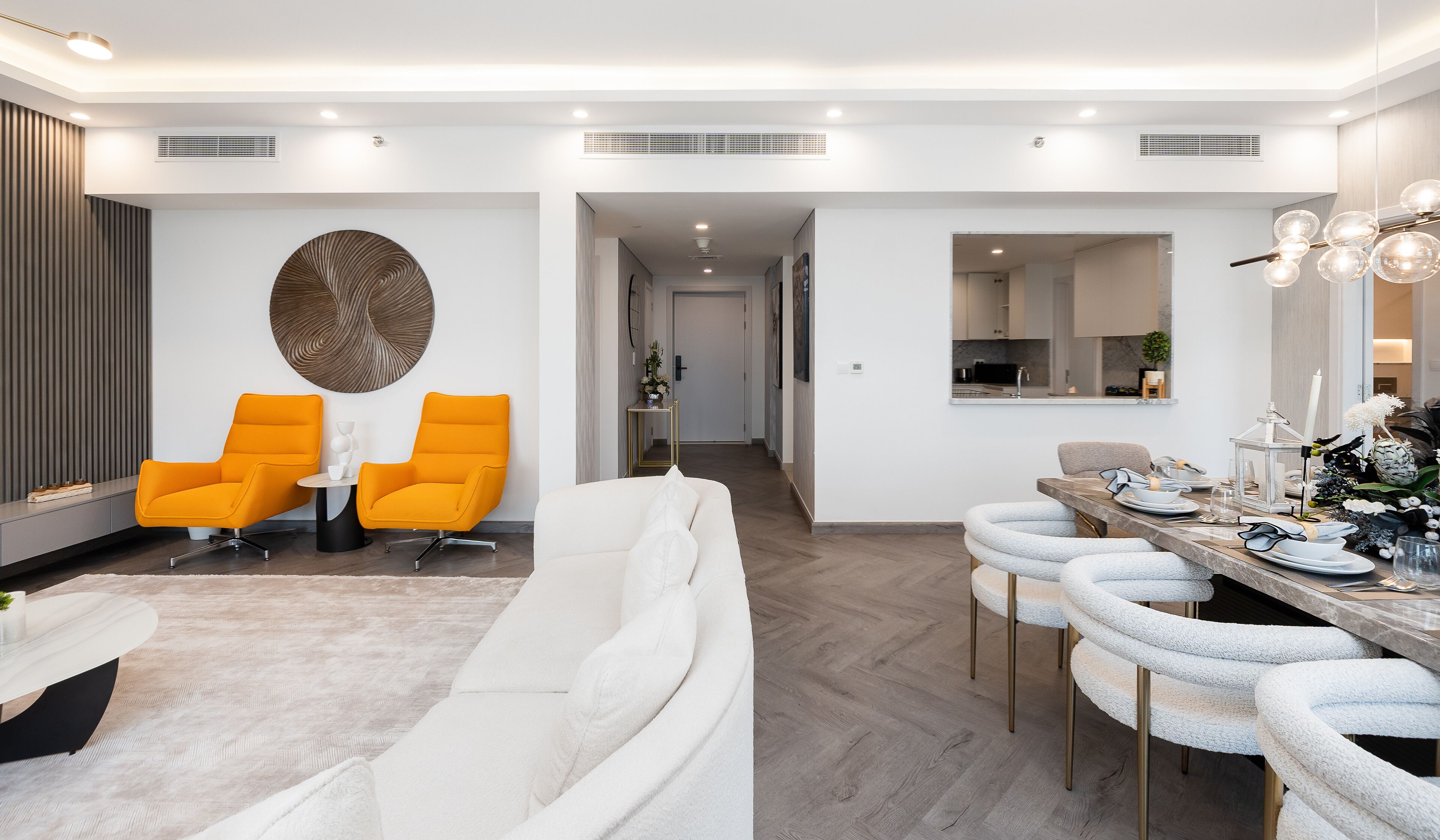 A modern living room with a white sofa, two orange armchairs, and a dining area with a marble countertop and white bar stools.