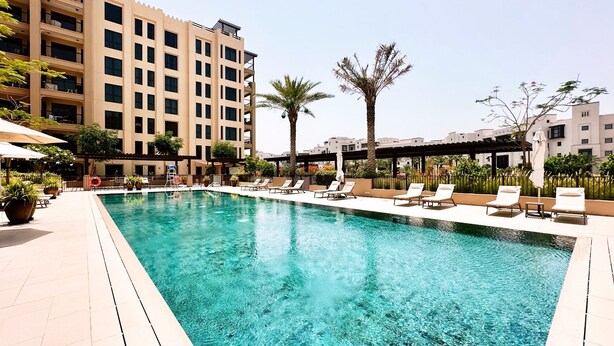 A swimming pool with lounge chairs and palm trees, surrounded by a multi-story building. - Lamtara 1 - Mint Stay (Dubai)