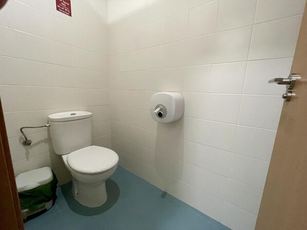 Un baño pequeño con azulejos blancos, un inodoro, dispensador de toallas de papel y un cubo de basura verde.