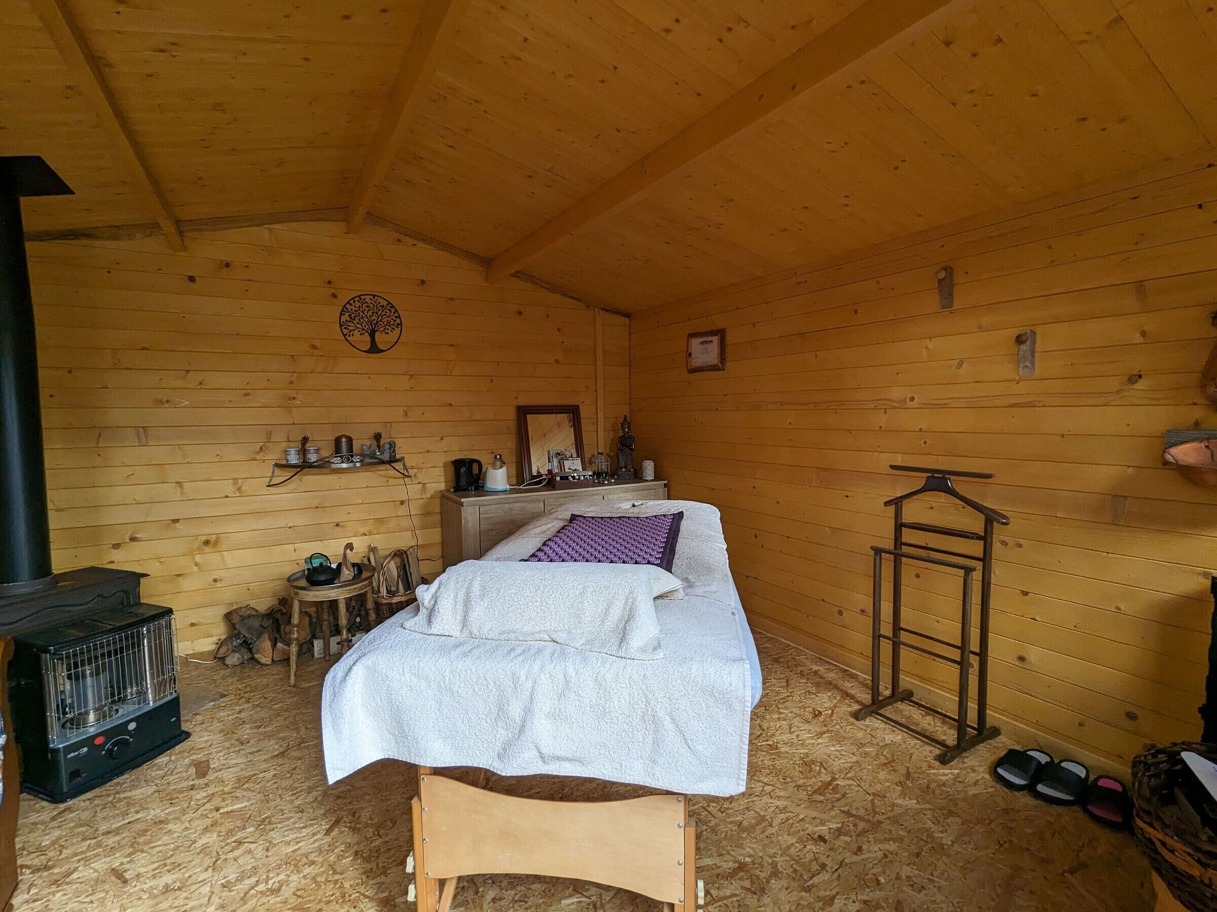 Acogedora habitación tipo cabaña de madera, con cama, calefactor, una mesita con objetos y una estantería con diversos artículos.