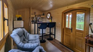 L’intérieur chaleureux d’un chalet avec un plafond en bois, un coin repas avec deux tabourets de bar, une cuisine équipée d’un évier et d’une cuisinière, et une grande fenêtre offrant une vue sur un paysage verdoyant.