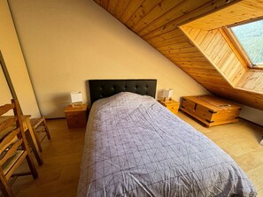 Ett sovrum med ett sluttande tak, en säng med ett mönstrat överkast, ett träbord som sängbord, en stol och en liten trälåda med lådor.