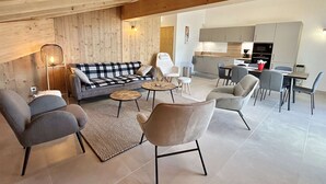 Una sala de estar moderna con un sofá, sillas y una mesa de centro. Se ve una cocina al fondo.