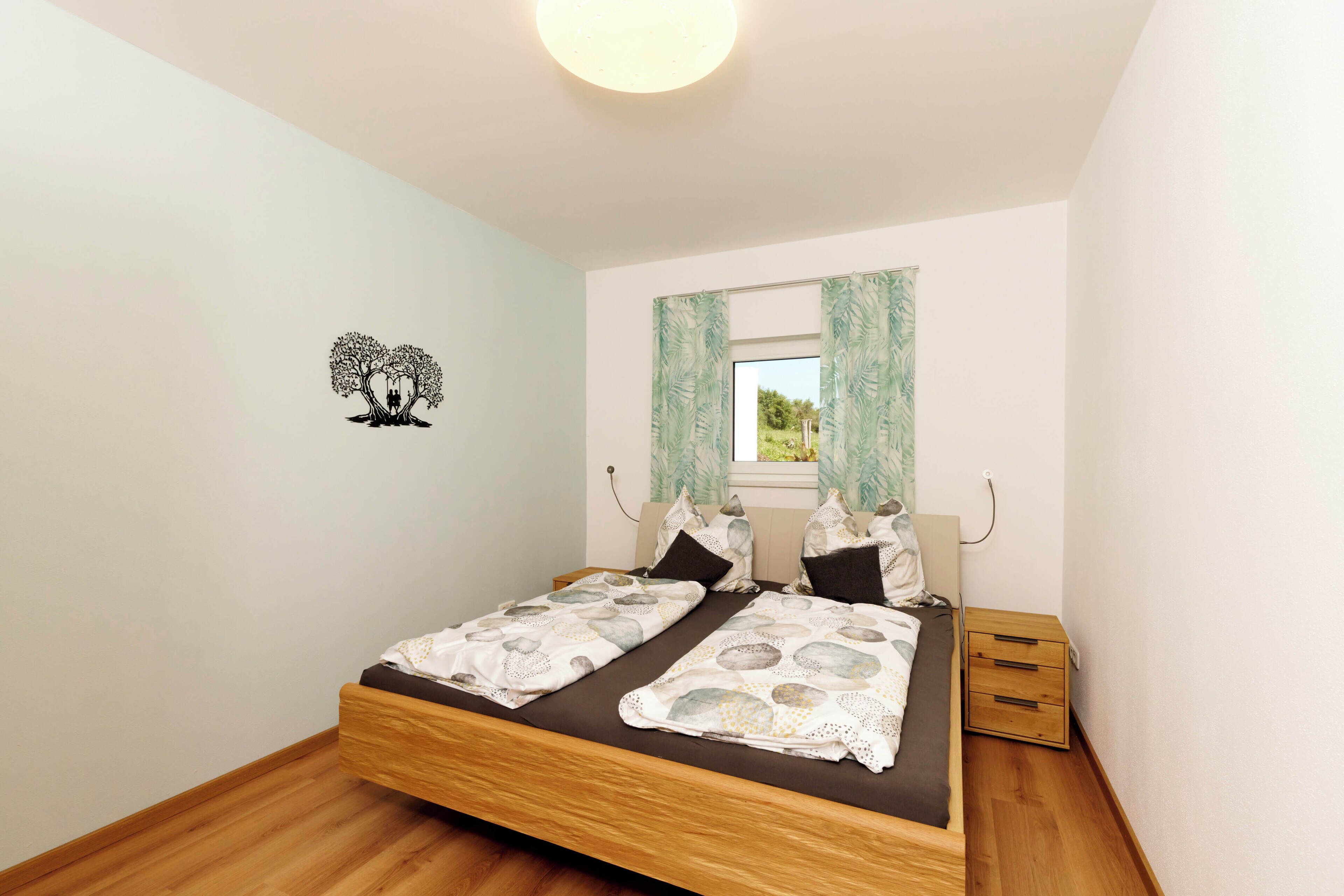 A double bed with a wooden frame, two bedside tables, a window with curtains, and a wall decal of a tree.