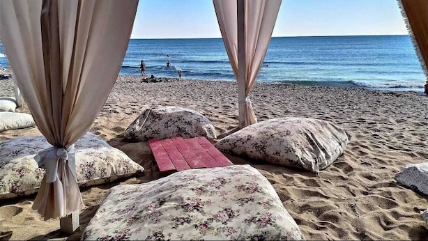 Blick auf einen Strand mit einer Holzbank und Kissen, Vorhängen und einem klaren Himmel.