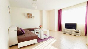 Ein modernes Wohnzimmer mit einem Ecksofa, einem weißen Couchtisch, einem Flachbildfernseher auf einem weißen Schrank und einer kleinen Regaleinheit.