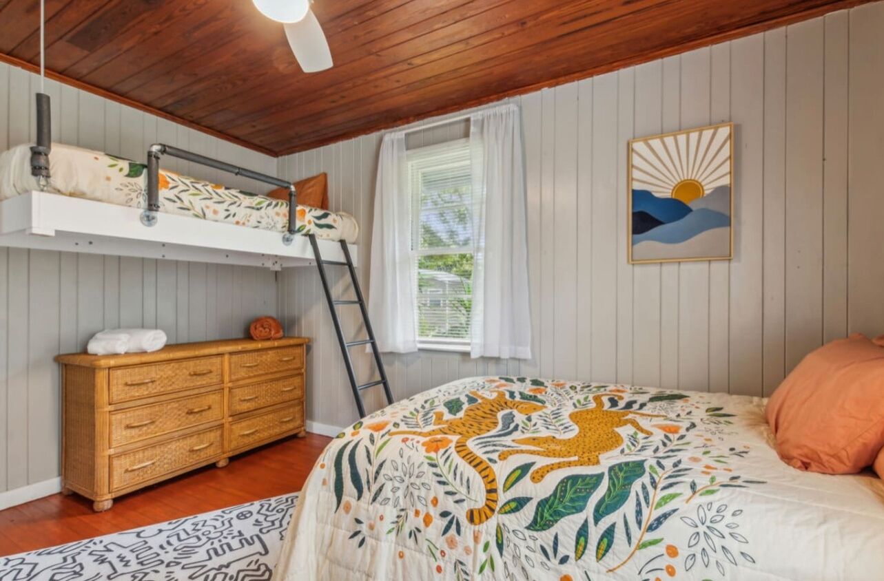 Une chambre avec un lit superposé, une commode en bois, une échelle, une fenêtre avec des rideaux blancs et un tableau accroché au mur.