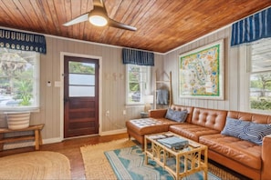 A cozy living room with a wooden ceiling, a brown leather sofa, a wooden coffee table, and a large painting on the wall. - Oak Grove Cottage at Fernandina Beach (Fernandina Beach)