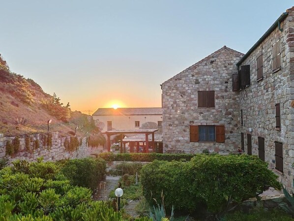 A stone building with a sloped roof, surrounded by greenery and a clear sky. - House on the Beach (Sardegna)