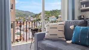 A balcony with a view of a hillside town, a grey sofa with decorative pillows, and a small table with a coffee maker. - Cozy Retreat in Romantic Zone - Gym & BBQ (Puerto Vallarta)