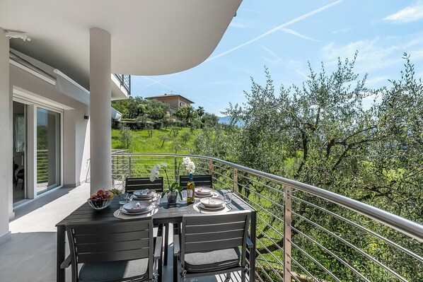A balcony with a table set for dining, overlooking a lush garden and a distant building. - Casa Beatrice (Tenno)