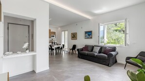 A modern living room with a grey sofa, a dining area with a white table and chairs, and a kitchen in the background. - Casa Beatrice (Tenno)