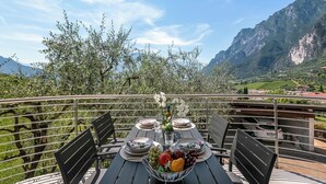 A table set with plates, a bowl of fruit, and a vase of flowers on a balcony overlooking a mountainous landscape. - Casa Beatrice (Tenno)