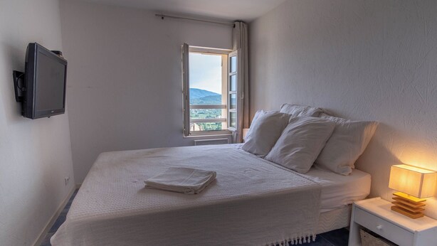 A neatly made bed with a white blanket and pillows, a bedside table with a lamp, and a TV mounted on the wall. - Duplexe with panoramic south-facing terrace in the center of Grimaud (Grimaud)