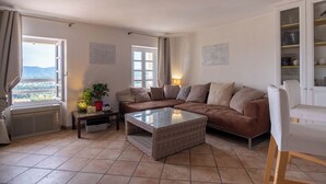 A living room with a sectional sofa, a glass-top coffee table, a white armchair, and a cabinet with shelves. - Duplexe with panoramic south-facing terrace in the center of Grimaud (Grimaud)