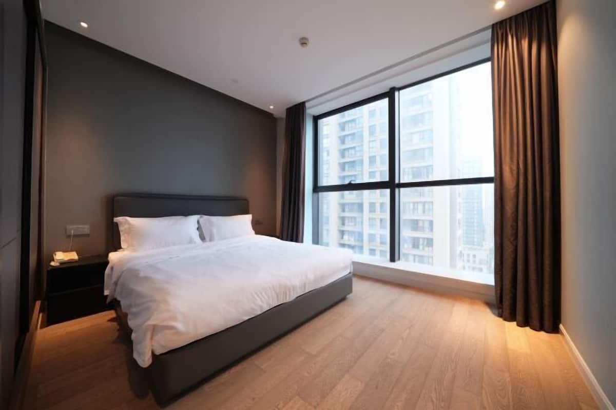 A modern hotel room with a large bed, a nightstand, a window with curtains, and a view of high-rise buildings.