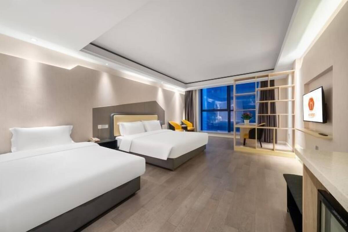 A modern hotel room with two beds, a TV, a desk, and a large window with a city view.