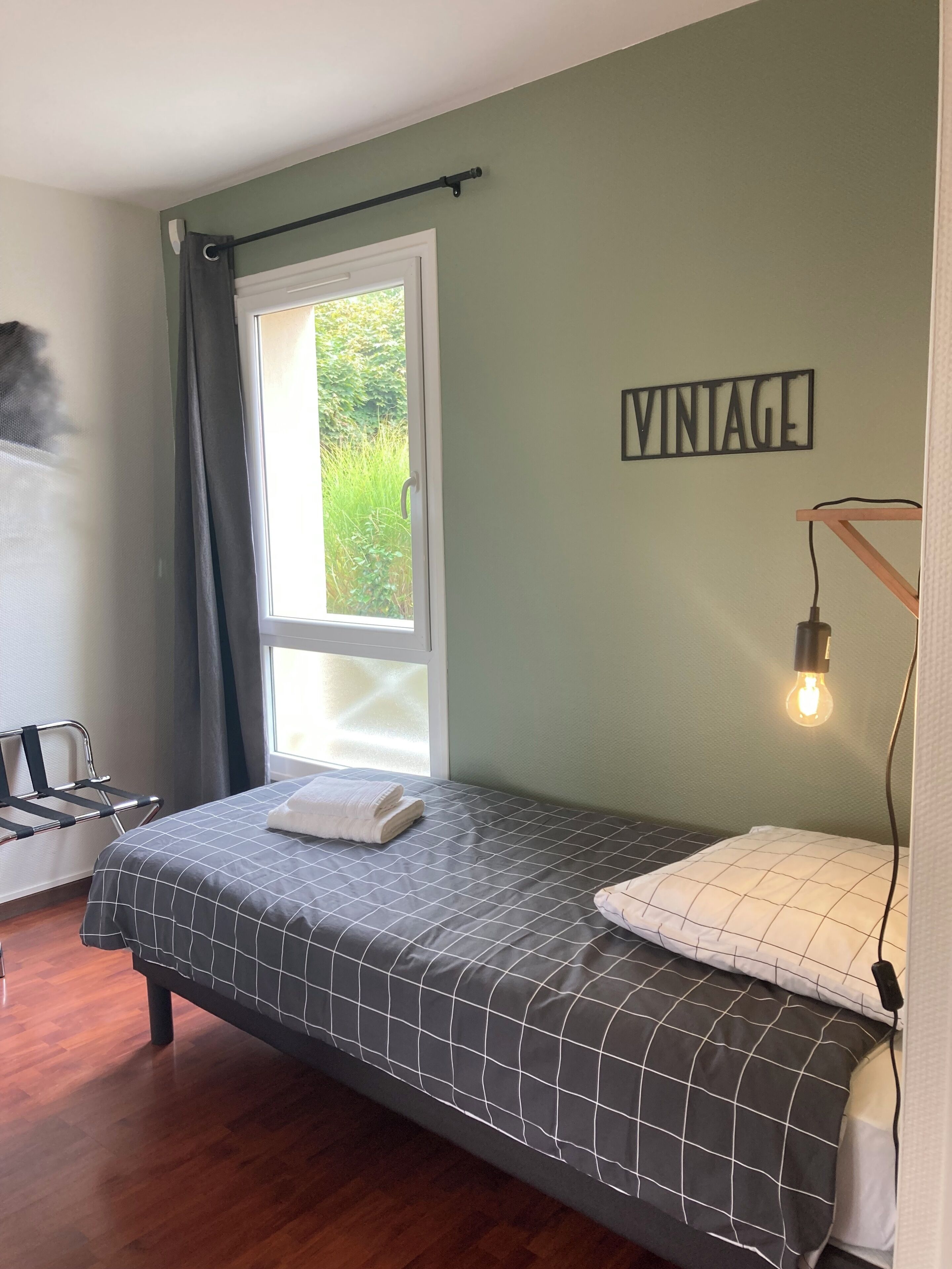 A small, neatly arranged bedroom with a bed, a window with curtains, a wall-mounted light, and a 'VINTAGE' sign.