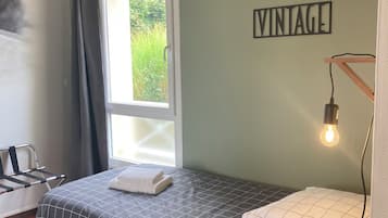 A small, neatly arranged bedroom with a bed, a window with curtains, a wall-mounted light, and a 'VINTAGE' sign.