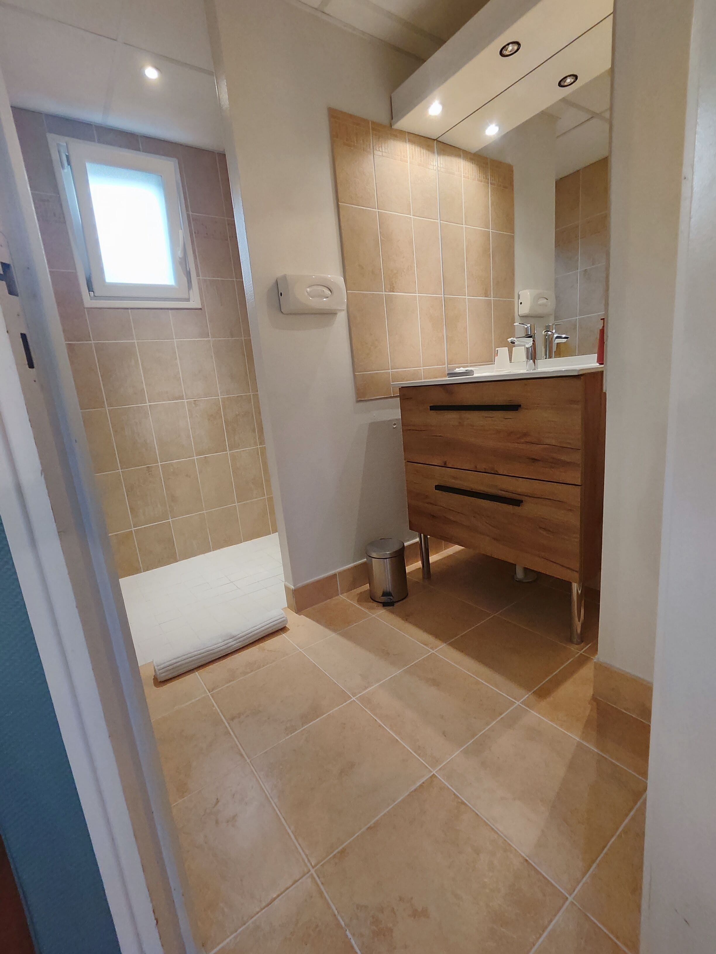 Ein Badezimmer mit beigen Fliesen, einem Holzkosmetikschrank, einem Spiegel und einem Fenster.